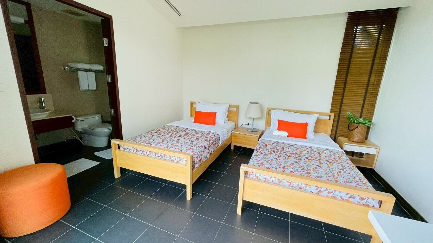 Een kamer met twee houten bedden, elk met een bloemenbedekking en oranje kussens. Er is een klein oranje krukje, een nachtkastje met een lamp, en een raam met jaloezieën.
