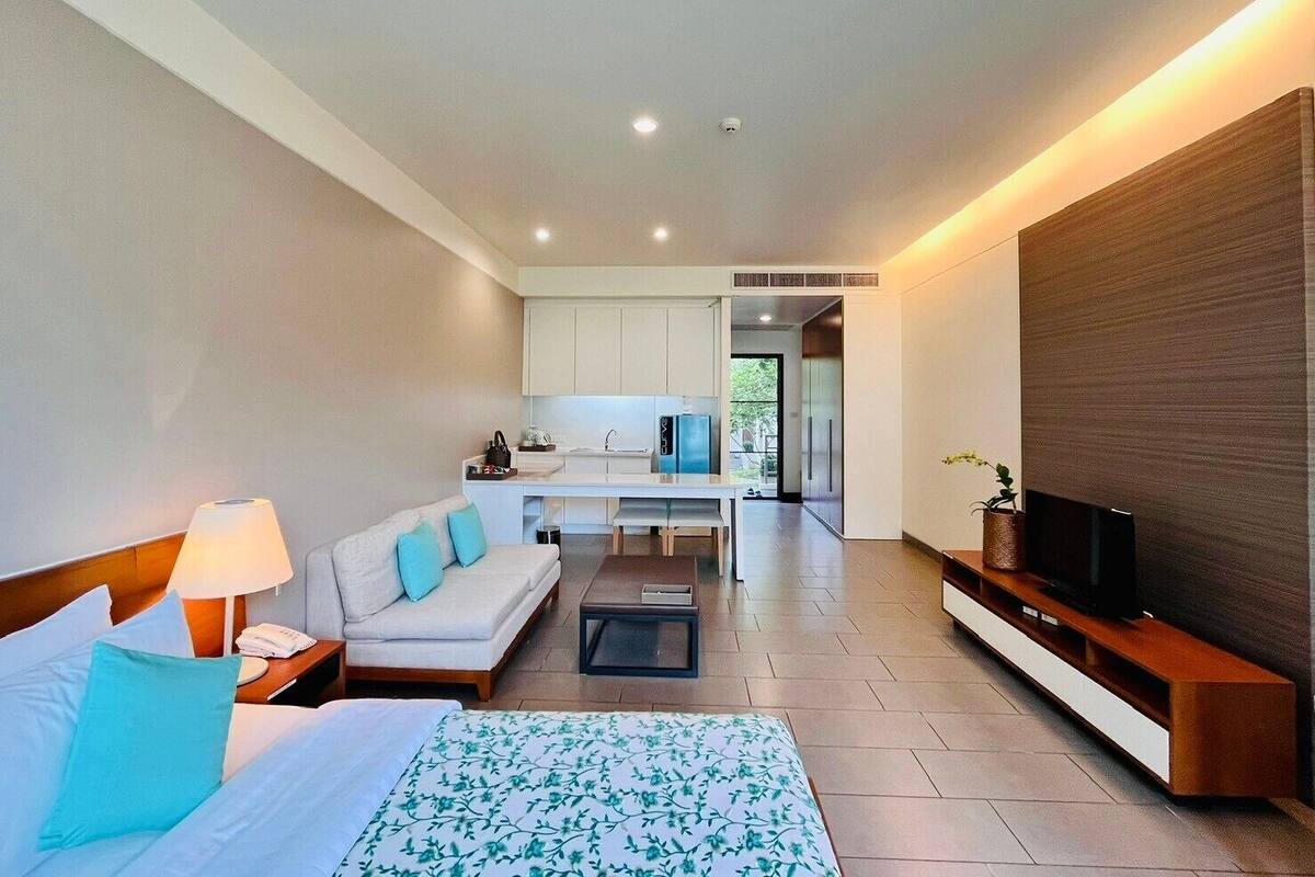 A modern hotel room with a bed, a sofa, a coffee table, a TV, a kitchen area, and a large window.