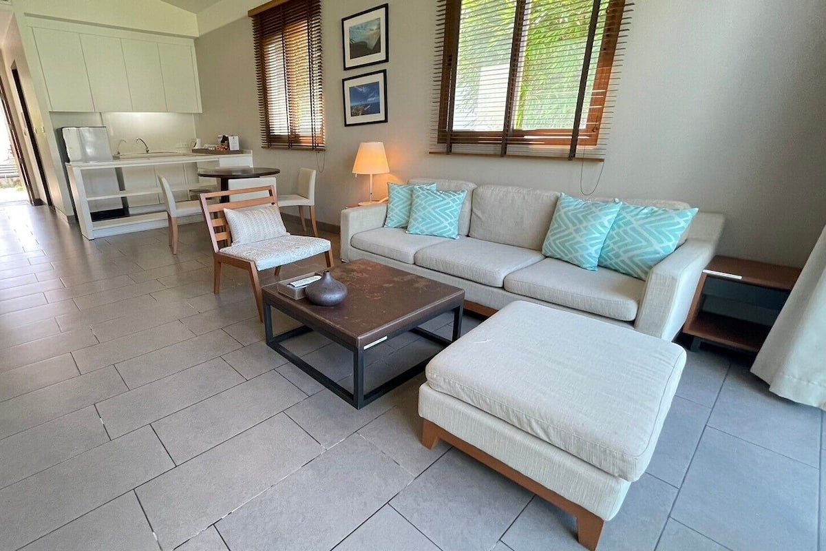 A spacious living area with a sofa set, a coffee table, and a dining table with chairs. There is a kitchen area in the background, and the room has large windows with blinds.
