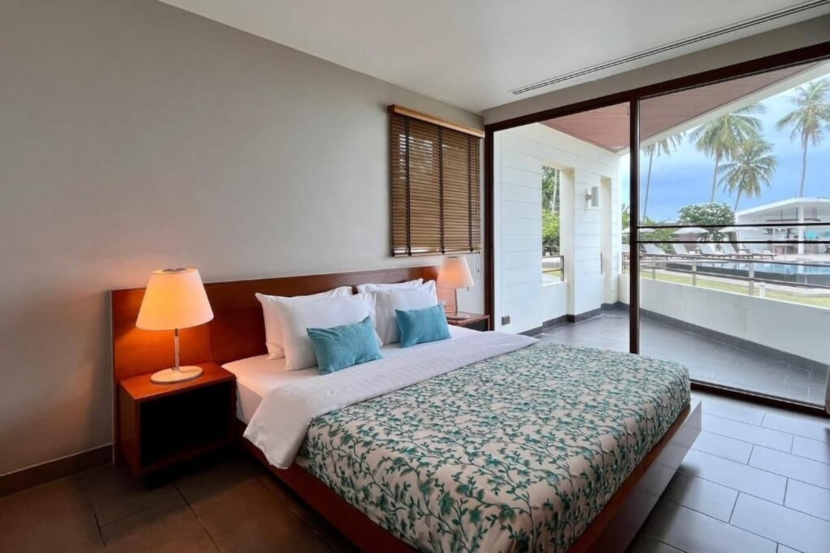 A hotel room with a large bed, a nightstand with a lamp, a sliding glass door to a balcony with a view of palm trees.
