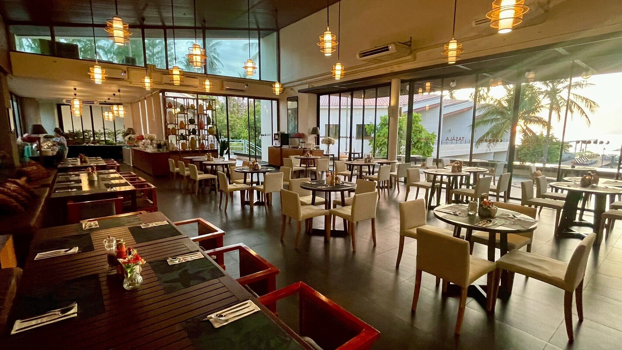 A spacious restaurant with multiple dining areas, large windows offering views of palm trees, and hanging pendant lights.