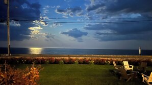 Un espace extérieur en bord de mer, avec une pelouse verdoyante, un coin salon avec des chaises et une vue sur la mer sous un ciel nuageux.