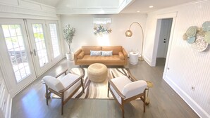 Un salon spacieux avec un plafond haut, une grande fenêtre, un canapé marron, deux fauteuils blancs, une table basse ronde et un lampadaire.