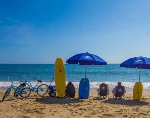 A beach scene with surfboards, bicycles, and beach chairs under umbrellas. - 7076 JMJ Seascape * Oceanfront * Pool * Hot Tub (Nags Head)