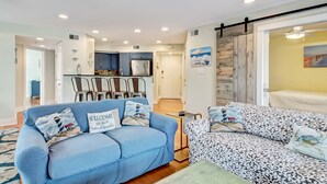 A modern living room with a blue sofa, patterned sofa, and a coffee table. There is a kitchen area with blue cabinets and a refrigerator. - Heated Pool | 5*Clean | Flex Cancelation (Tybee Island)