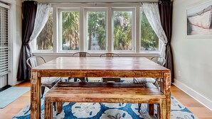 A dining area with a wooden table and benches, large windows with curtains, a ceiling fan, and a patterned rug. - Heated Pool | 5*Clean | Flex Cancelation (Tybee Island)