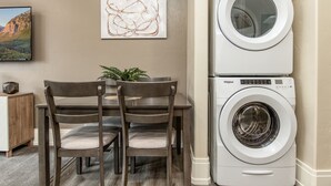 A laundry room with a stacked washer and dryer, a dining table with chairs, and a framed abstract painting on the wall.