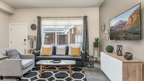 A modern living room with a sofa, armchair, and coffee table, a large window with curtains, a flat-screen TV, and a view of a mountain landscape.