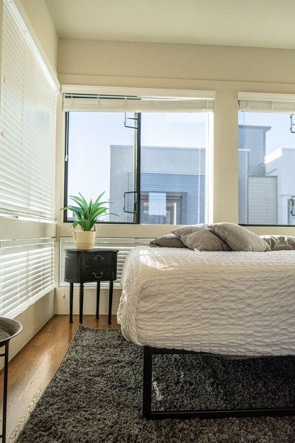 Une chambre à coucher avec un lit, une petite table sur laquelle est posée une plante, et une vue sur les immeubles à travers de grandes fenêtres.