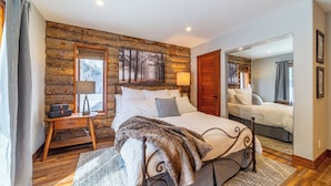 Een slaapkamer met een wand van houten boomstammen, een groot bed met metalen frame, een nachtkastje met lamp en een spiegel aan de muur.