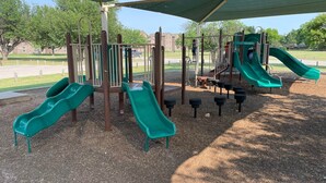 A playground with multiple slides and climbing structures, surrounded by a covered area and trees.