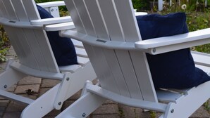 Deux chaises Adirondack blanches, dotées de coussins foncés, sont placées sur une zone pavée, offrant une vue sur une route et sur la verdure en arrière-plan.