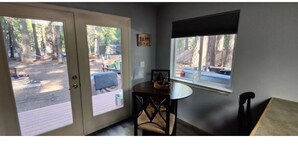 A room with a door leading to a deck, a small table with a chair, and a window with a view of trees. - Mt Lassen and the great outdoors (Shingletown)