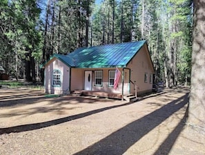 Situated in a forested area, this cabin features a front porch with a bench and flagpole.