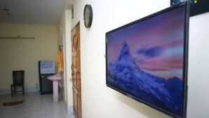 A hallway with a large TV screen displaying a mountain landscape, a wooden door, a small desk with a chair, and a tiled floor. - 💗AC,ATTACH BATH ROOM,NEAR LALMATIA MOHAMADPUR (Dhaka)