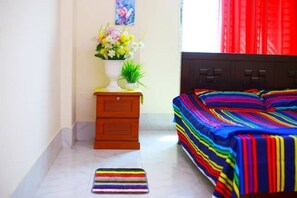 A bedroom with a bed covered in a multicolored striped blanket, a small wooden nightstand with a white vase of flowers, and a striped rug on the floor. - 🏵️CUTE ROOM,MOHAMMADPUR LALMATIA (Dhaka)