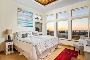 Quarto com uma cama grande, mesa de cabeceira, escrivaninha e várias janelas com vista para a cidade.