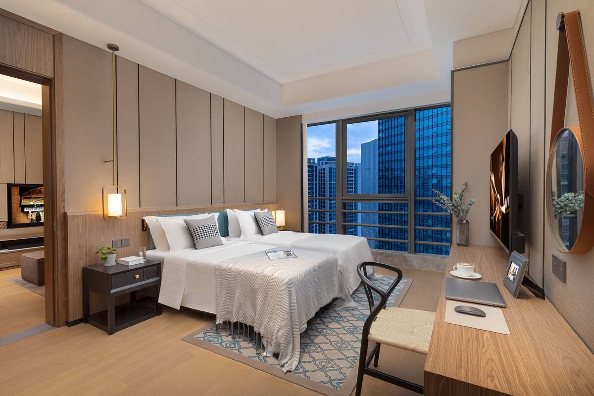 A modern hotel room with a large bed, a desk with a laptop, a chair, a TV, and a view of the cityscape through large windows.