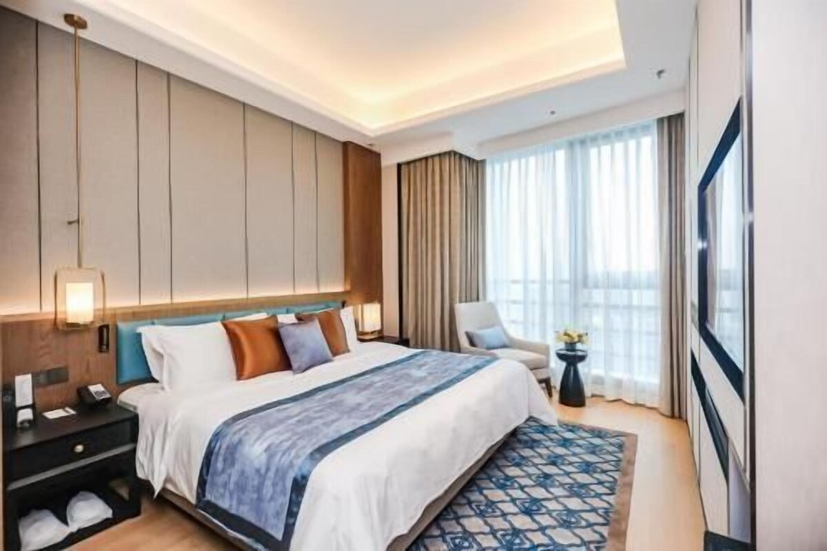 A modern hotel room with a large bed, a bedside table, a chair, and a view of the city.