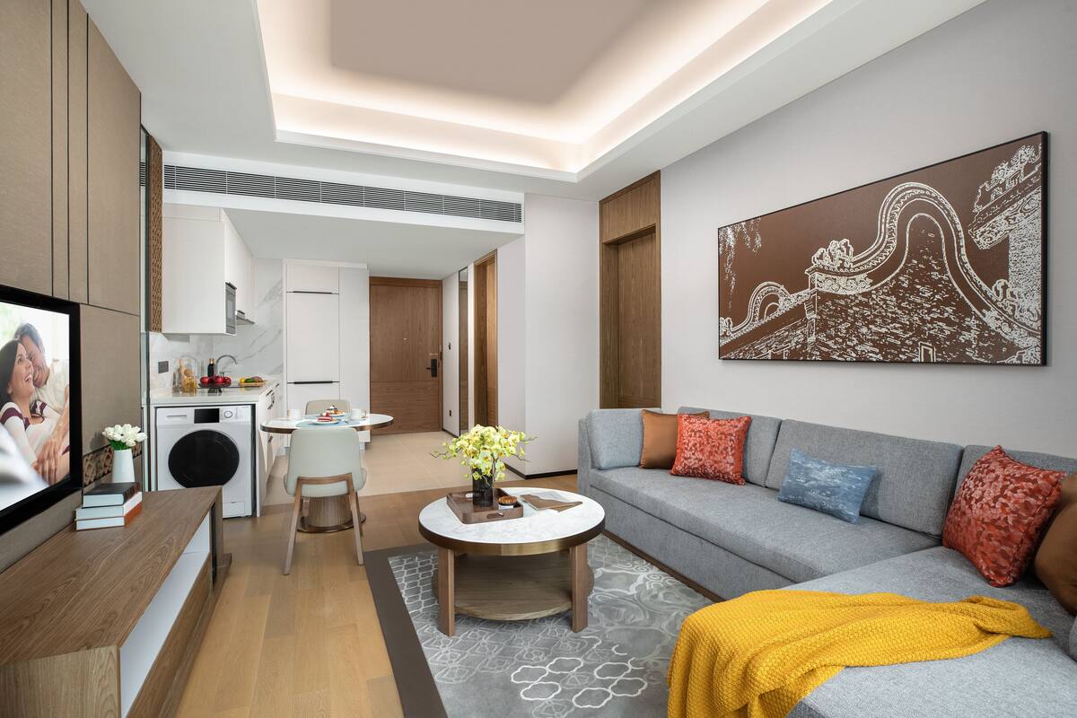 A modern living room with a grey sofa, a coffee table, and a TV. There is a kitchen in the background with a washing machine and a dining area.