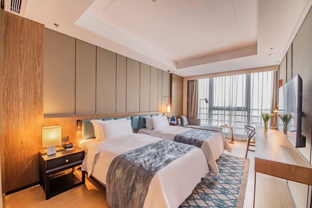 A modern hotel room with two beds, a desk, a TV, and a large window.