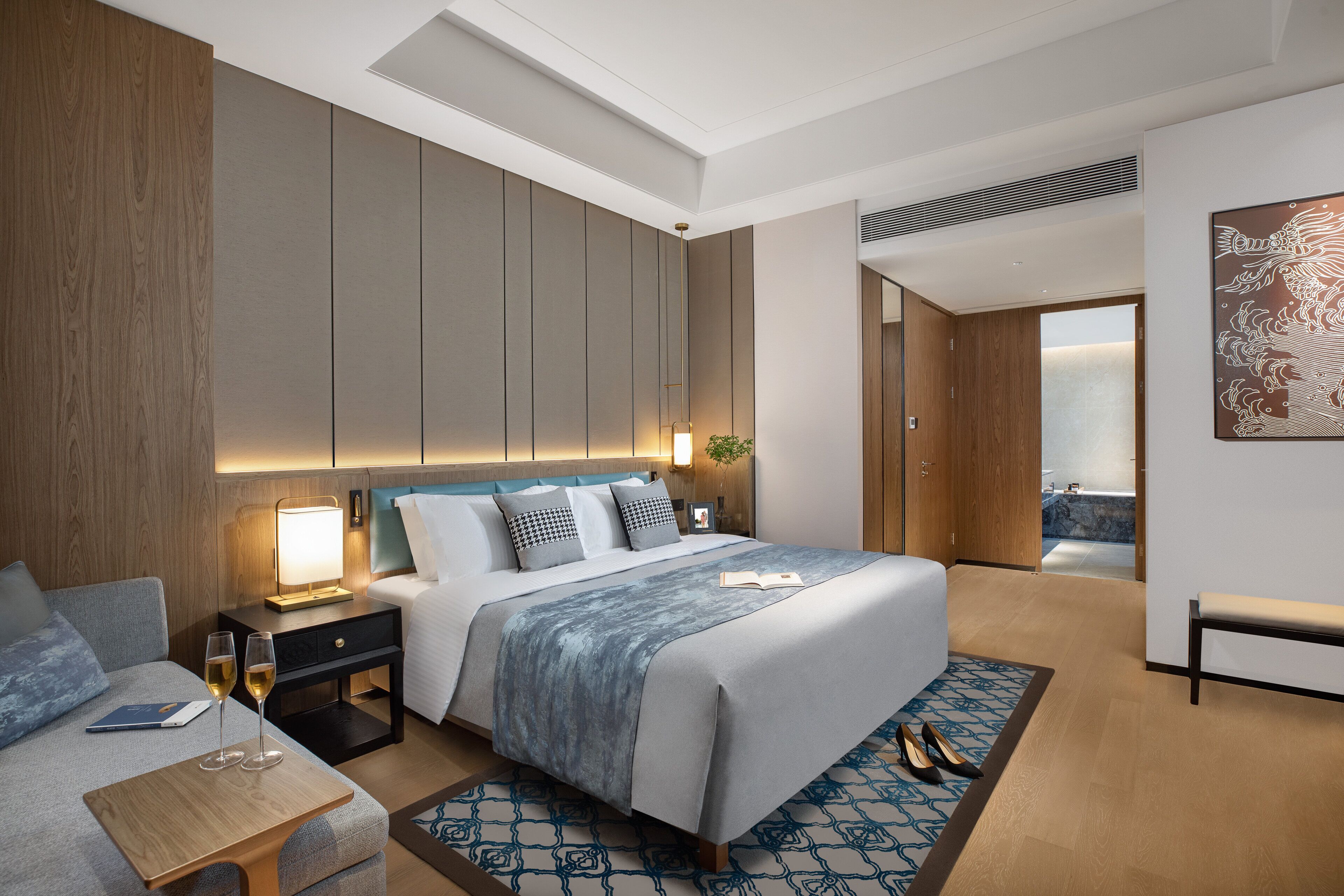 Una habitación de hotel moderna con una cama grande, una mesita de noche con lámpara, un área de descanso y vistas a un balcón.