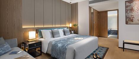 A modern hotel room with a large bed, a bedside table with a lamp, a small seating area, and a view of a balcony.