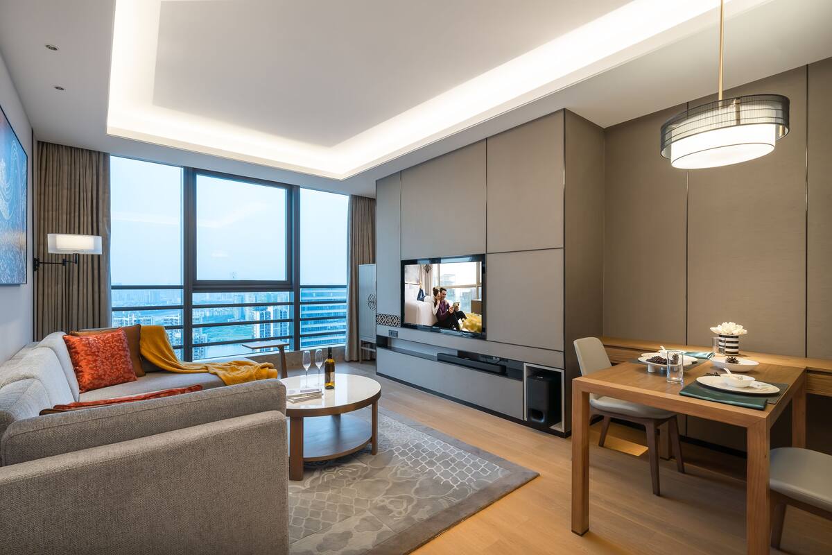 A modern living room with a sofa, coffee table, and dining area. A television is mounted on the wall, and there is a large window providing a view of the city.