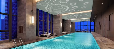 An indoor swimming pool with tiled flooring, lounge chairs, and a ceiling with decorative lighting.