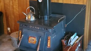Een Blaze King houtkachel met een waterkoker erop, omringd door houtblokken en een metalen pot.