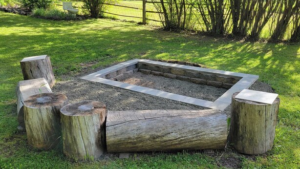 Een vuurkorf met een rechthoekige stenen rand, omgeven door houten palen, gelegen in een grasveld met een hek en bomen op de achtergrond.