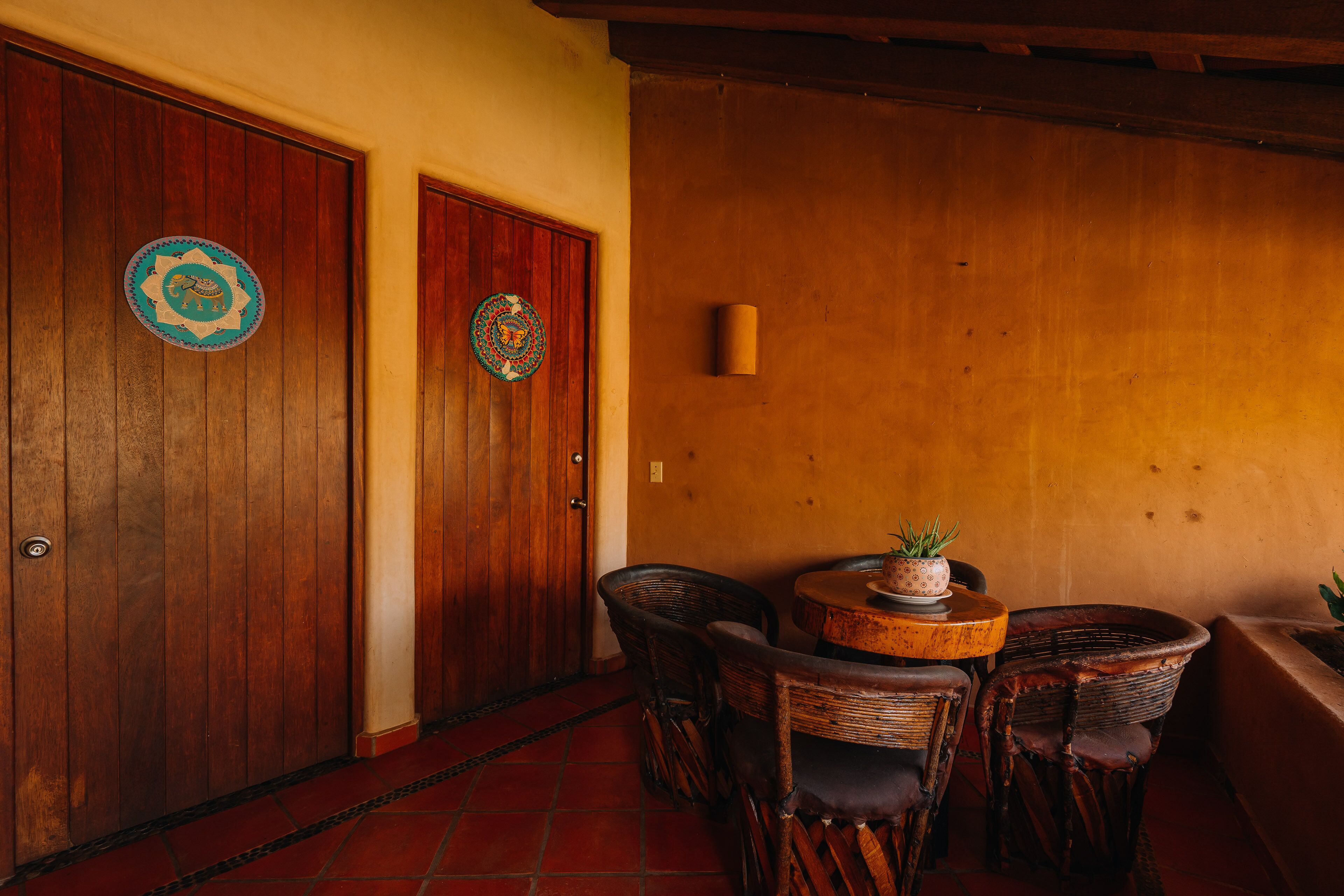 A wooden door with a circular emblem, a small table with a plant, wicker chairs, and a tiled floor.