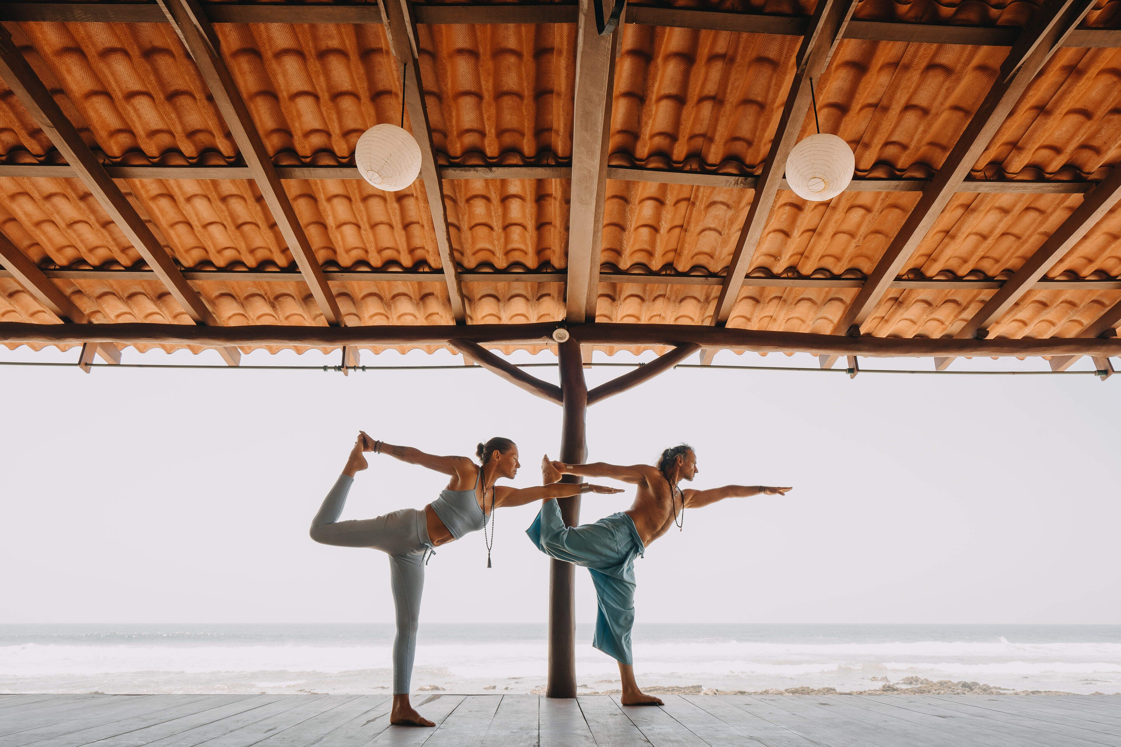 Two people practicing yoga under a wooden structure with a tiled roof by the beach.