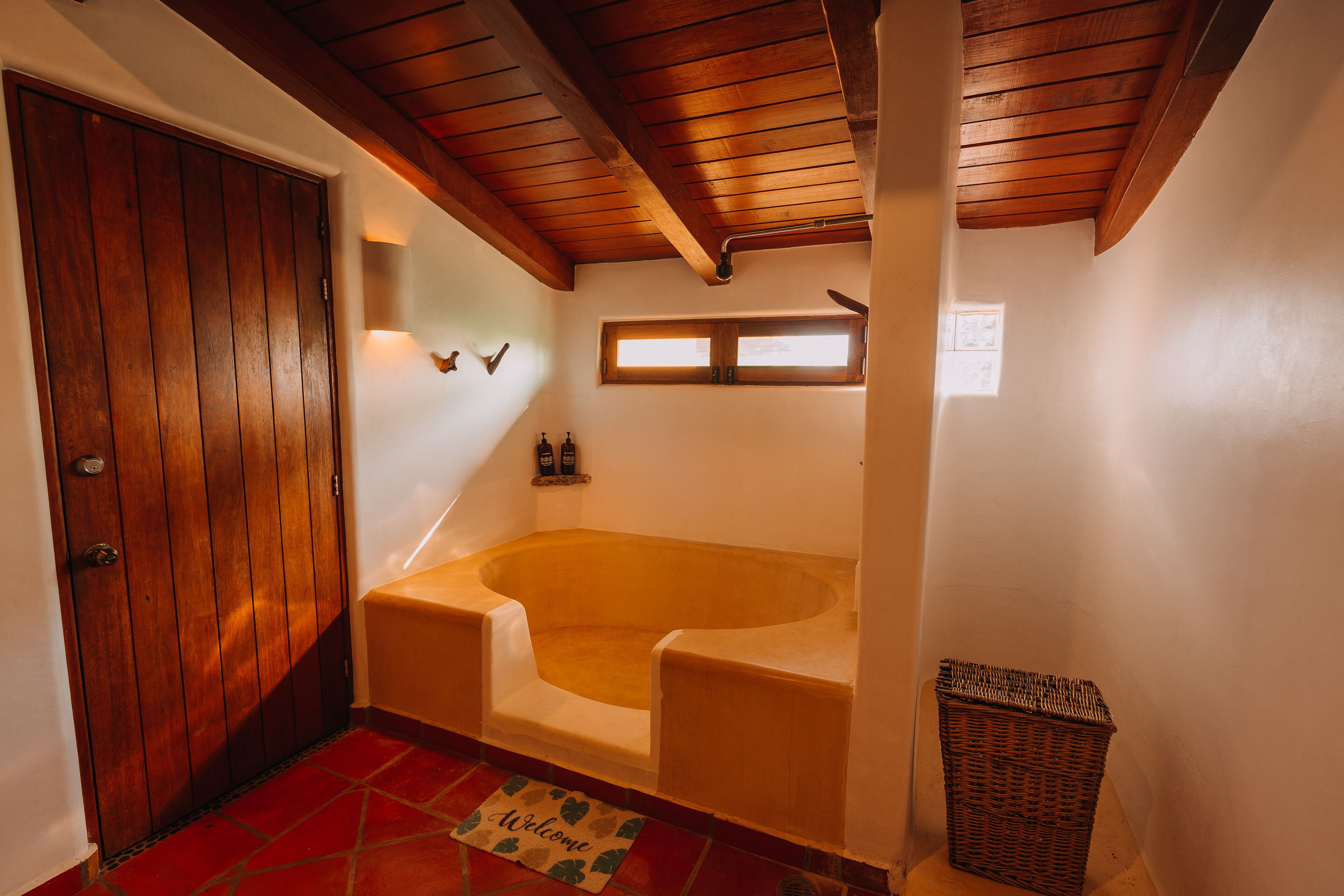 Un baño con puerta de madera, una bañera, una cesta de mimbre y un felpudo.