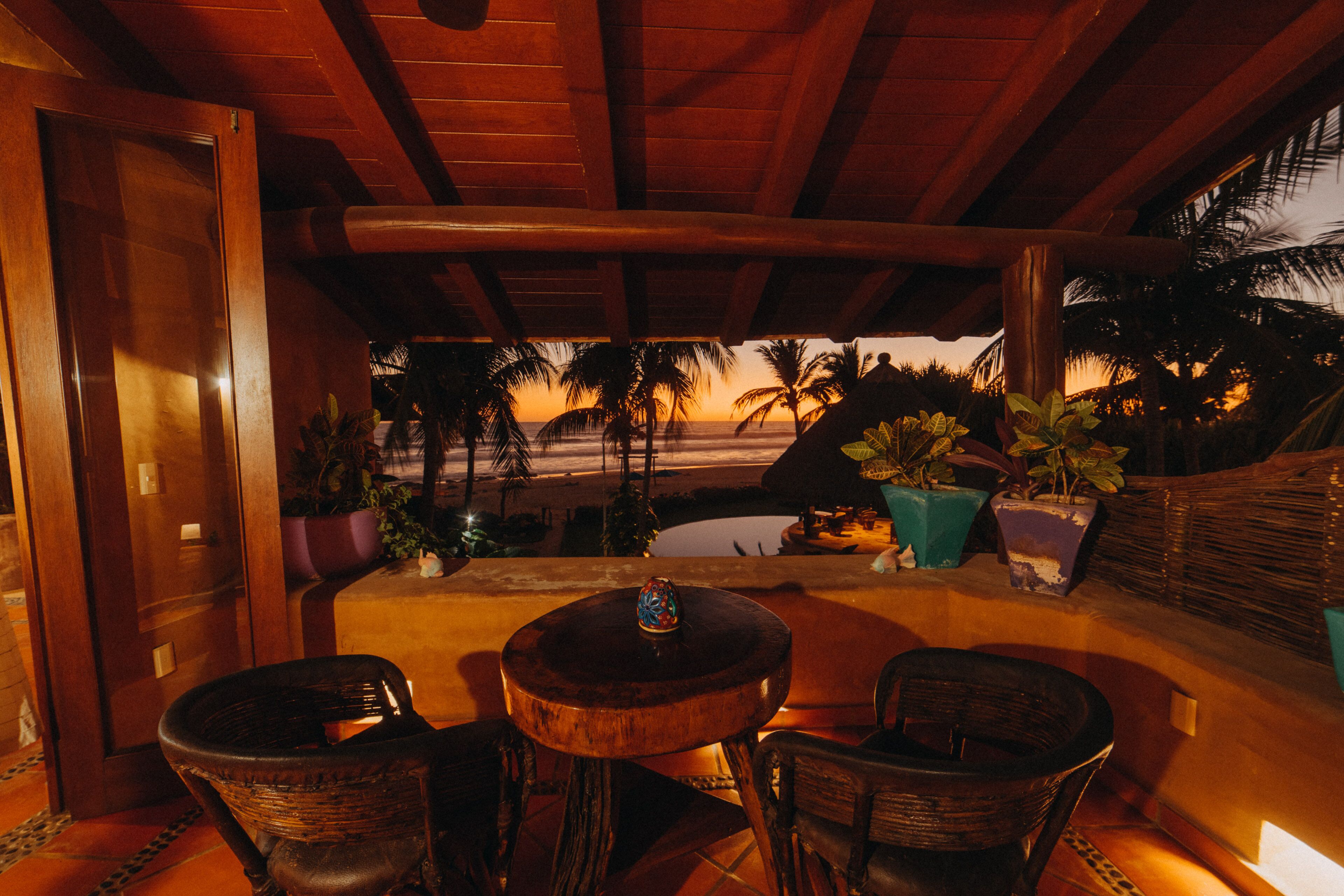 Un espace extérieur aménagé avec des chaises en osier et une table ronde, offrant une vue sur une plage bordée de palmiers et un coucher de soleil.