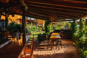 Un patio al aire libre con mesa y sillas de madera, rodeado de abundante vegetación y piso de ladrillo.