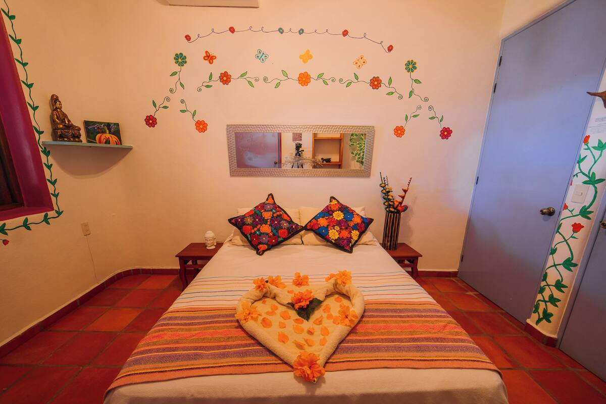 A neatly made bed with a heart-shaped pillow arrangement, a mirror above it, and a floral wall decoration.