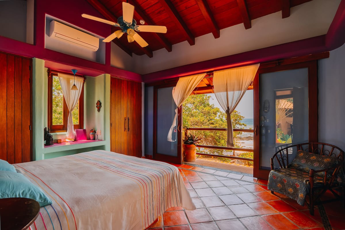 A bedroom with a bed, a chair, a window with curtains, a ceiling fan, and a view of the outdoors.