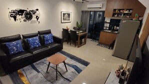 A living room with a black leather sofa, a wooden coffee table, and a dining area with a small table and chairs. There is a kitchen area with a refrigerator and cabinets.
