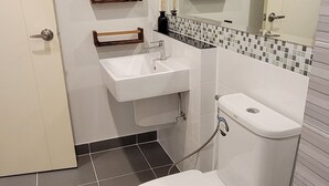 A bathroom with a toilet, sink, and mirror. There are wooden shelves above the sink and a decorative mat on the floor.