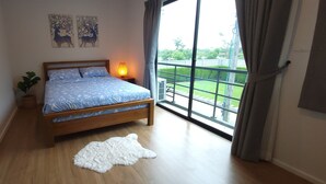 A bedroom with a wooden bed, a white furry rug, a nightstand with a lamp, and a balcony with a view of greenery.