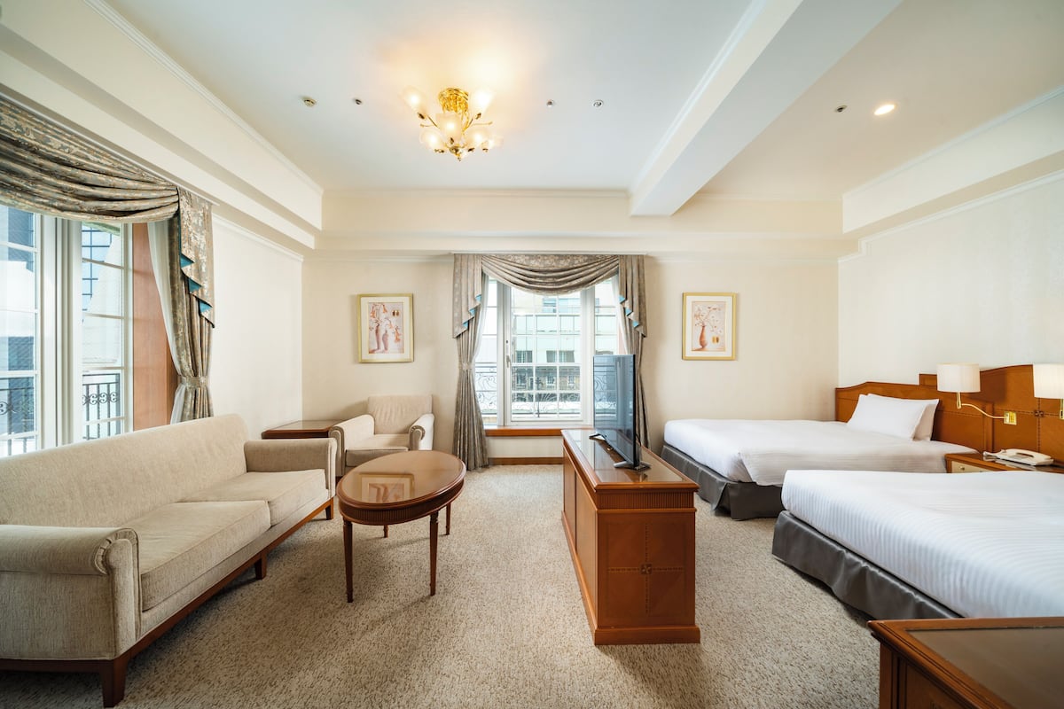 Une chambre d’hôtel avec un canapé, un bureau, deux lits, une télévision et une fenêtre avec des rideaux.