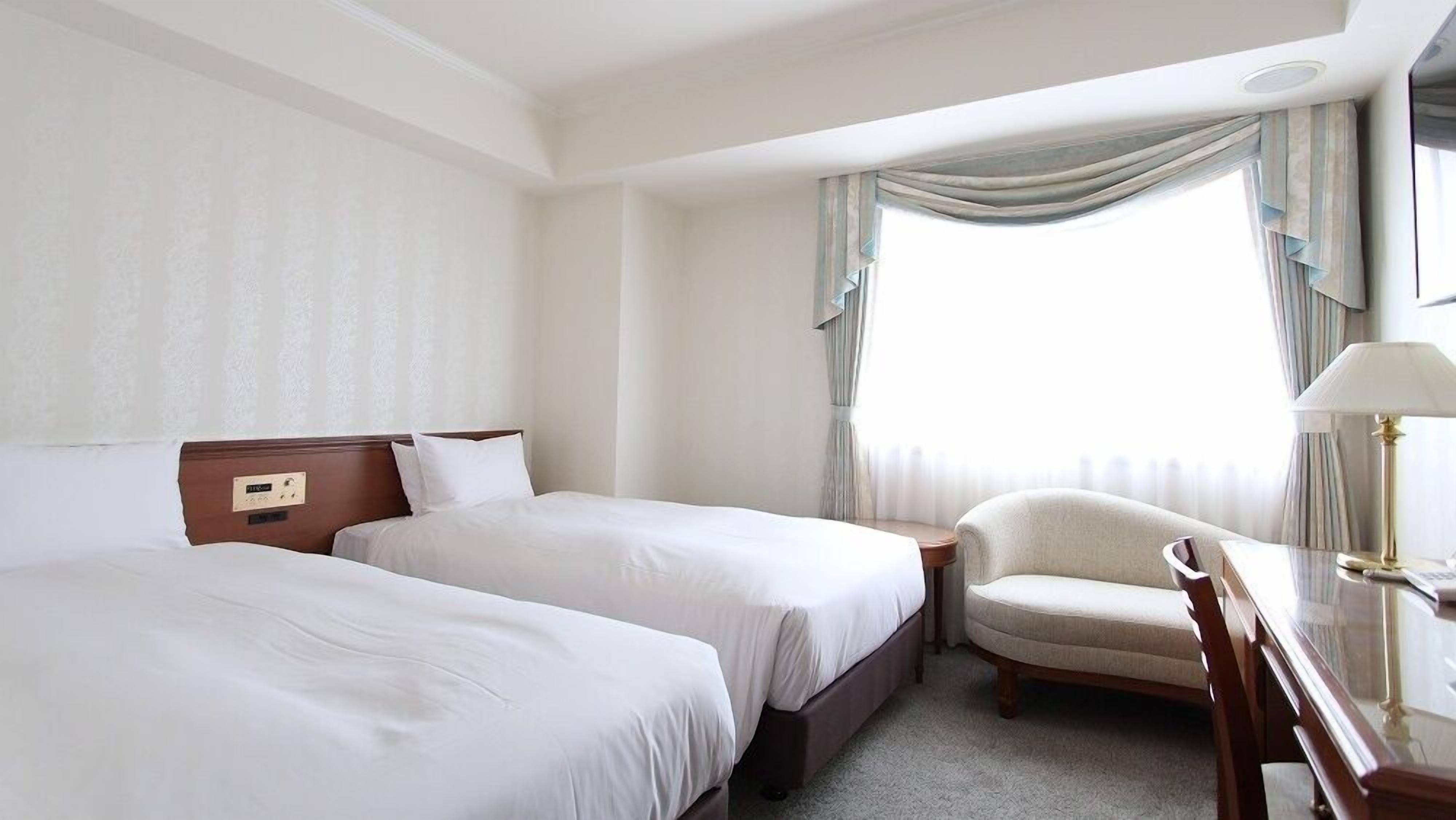 A hotel room with two beds, a chair, a desk, and a window with curtains.