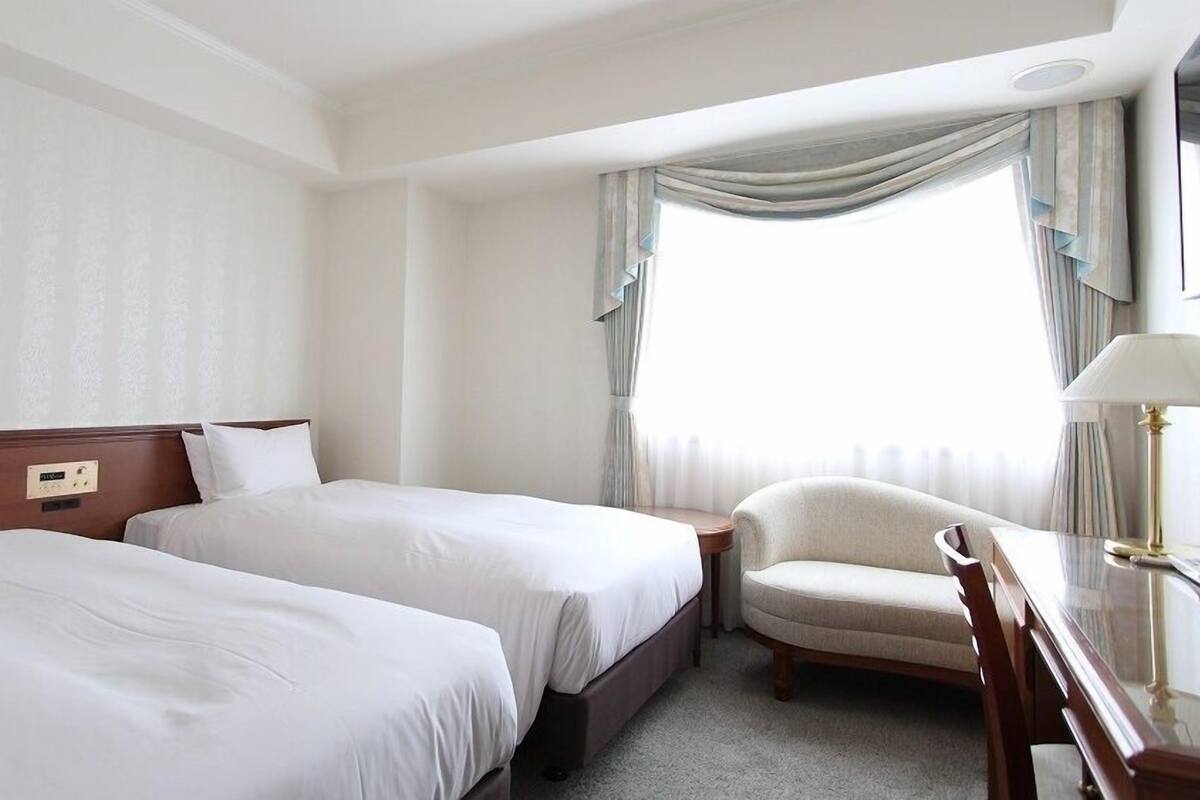 Une chambre d’hôtel avec deux lits, une chaise, un bureau et une fenêtre avec des rideaux.