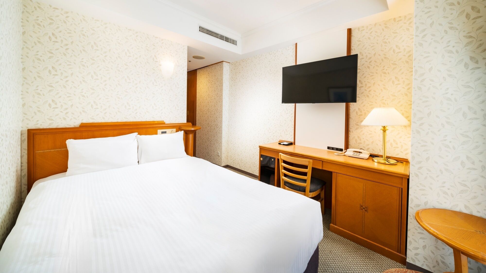 A hotel room with a bed, a desk, a chair, a TV, and a lamp.