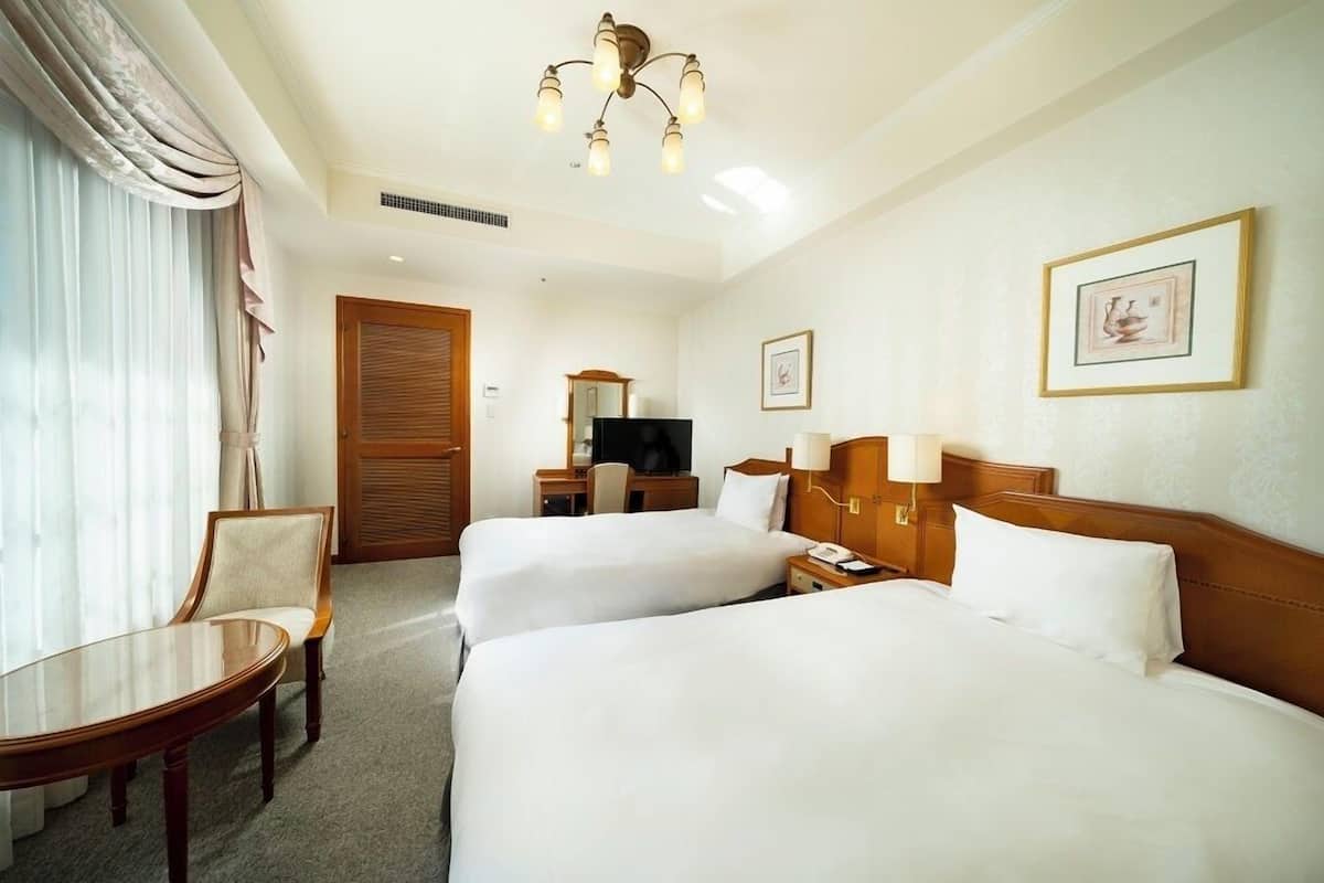 Une chambre d’hôtel avec deux lits, un bureau, une chaise, une télévision et un lustre.