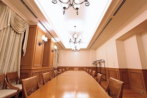Une salle de conférence avec une longue table, des chaises en bois, des lustres et une grande fenêtre avec des rideaux.