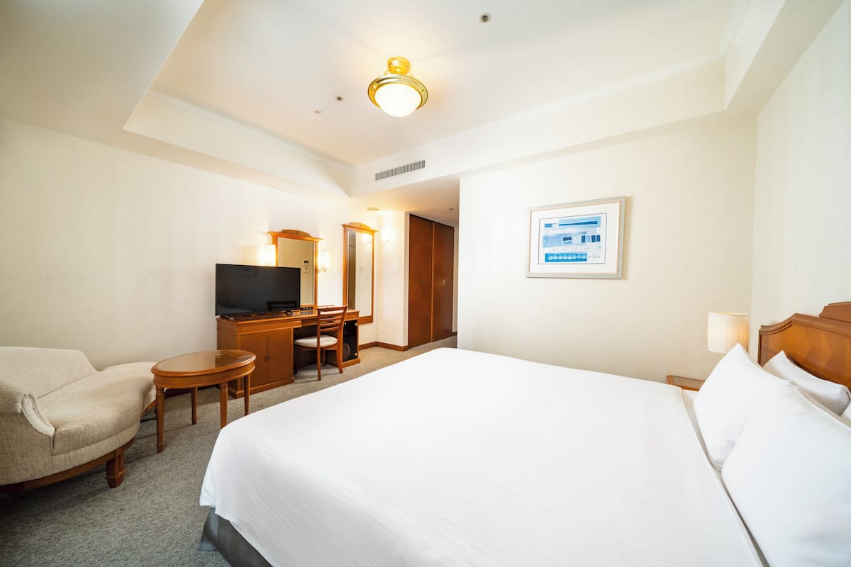 Une chambre d’hôtel avec un grand lit, un bureau avec une chaise, une télévision, une armoire et un tableau encadré au mur.