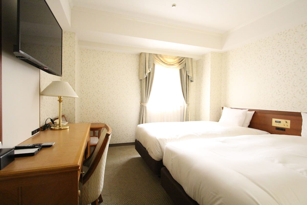 Une chambre d’hôtel avec deux lits, un bureau avec une lampe, un téléviseur fixé au mur et une fenêtre avec des rideaux.