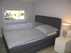 Una cama bien hecha con cabecera gris y ropa de cama blanca, una mesita de noche con lámpara, y una ventana con vistas al exterior.